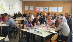 Four tables in a room with five or six people at each table. There are bottles of water on the near table and papers. The people are leaning in to talk with each other.