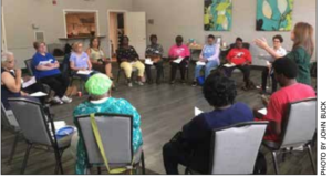 About twenty people sitting in a room in a circle of chairs. One woman is standing