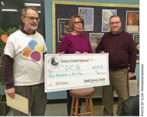 Three people holding a large simulated check. One man is wearing a white tee shirt with the CDCG logo